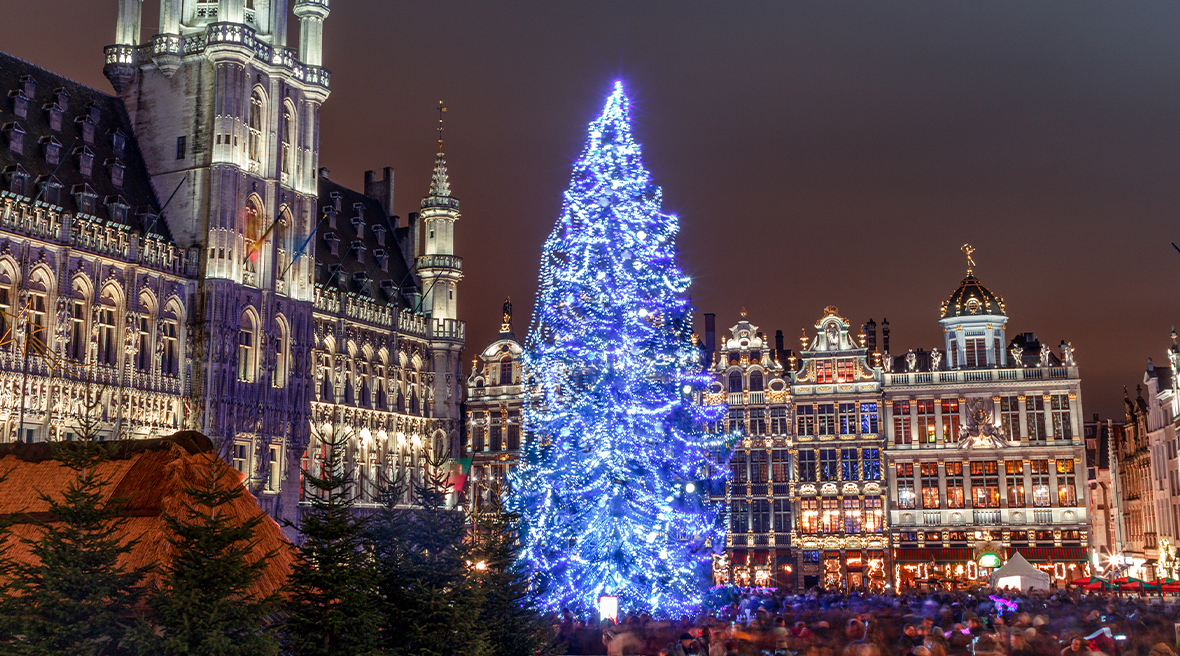 A Christmas tree lit up with blue lights surrounded by tall Grand buildings and a crowd below